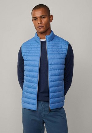 Chaleco acolchado sin mangas de color azul claro, con textura de almohadillado, cuello alto y cremallera en la parte delantera. Lleva puesto un suéter de manga larga de color azul oscuro.