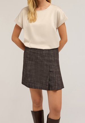 Crèmekleurige blouse met opgerolde mouwen, gecombineerd met een donker geruite minirok met een plooi aan de voorkant en een gladde textuur. Bruine laarzen tot aan de knieën.