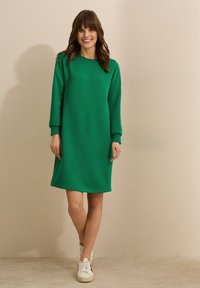 Grünes Sweatshirt-Kleid mit langen Ärmeln und gerippten Manschetten, ausgestattet mit Knopfakzenten an den Schultern. Der Stoff wirkt strukturiert und bequem.