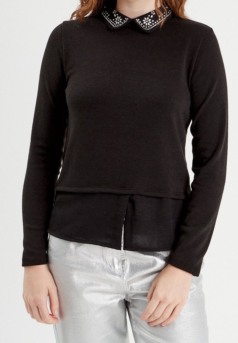 Femme portant un haut noir à manches longues avec un col décoratif orné de bijoux superposé à une chemise noire, associé à un pantalon métallique argenté.