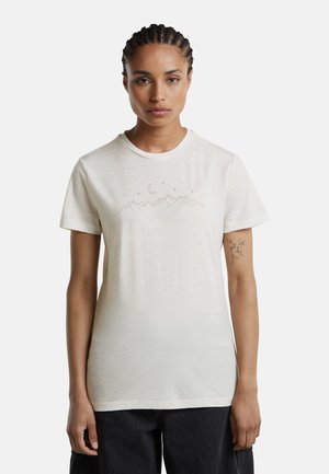 Frau mit geflochtenem Haar, die ein weißes T-Shirt mit einem Berg-, Mond- und Sternedesign trägt, steht vor einem schlichten hellen Hintergrund.