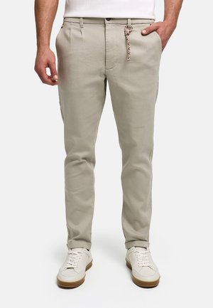Mężczyzna noszący beżowe spodnie slim fit z bocznymi kieszeniami, sznurkowy uchwyt pętli paska, białe sneakersy i białą koszulę włożoną do środka.