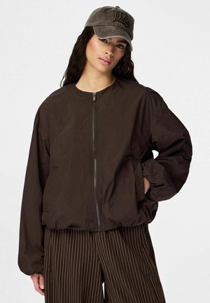 Nainen, jolla on päällään tummanruskea vetoketjullinen takki, ruskeat ohutraitat housut ja harmaa lippalakki, jossa on logo, poseeraa yhdellä kädellä taskussa.