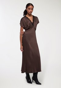 Abito in satin marrone con profondo scollo a V decorato con pizzo nero, maniche corte a dolman e una silhouette fluida fino alla caviglia. Stivali neri con tacco.