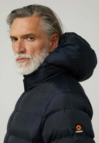 Abrigo acolchado azul marino con un cuello alto y una capucha desmontable, que presenta un acabado texturizado y un parche de logo naranja en la manga.