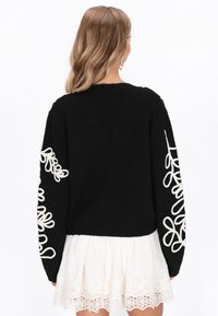 Čierny pletený sveter s krémovými vyšívanými kvetinovými vzormi na rukávoch. Krátky strih s rebrovaným lemom. Nosený cez bielu sukňu s čipkovým lemom.