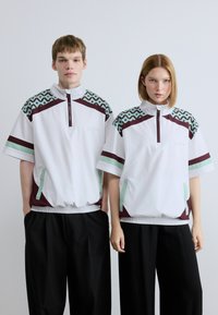 Πουλόβερ με σχεδίαση μισού φερμουάρ, λευκό υλικό, πολύχρωμο γεωμετρικό μοτίβο στους ώμους και ελαστικό τελείωμα· συνδυασμένο με μαύρο παντελόνι.