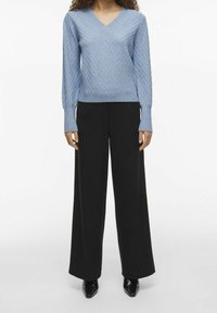 Pull en maille bleu clair avec col en V, motif chevron texturé ; associé à un pantalon noir à jambes larges et des chaussures noires à bout pointu.