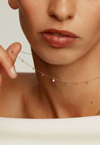 Collar de cadena de oro con pequeños colgantes circulares. Diseño delicado y textura suave, descansando sobre el cuello y sujetado por los dedos.