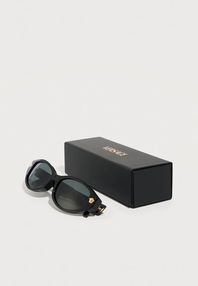Μαύρα γυαλιά ηλίου σχεδιαστή με σκούρους φακούς και χρυσές λεπτομέρειες δίπλα σε ένα μαύρο ορθογώνιο κουτί με χρυσό λογότυπο Versace.