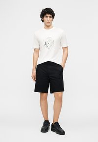 Young man with curly hair wearing a white graphic tee, black shorts, and black sneakers, standing with one hand in pocket.