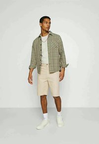 Ung man i en grön rutig skjorta över en vit t-shirt, beige shorts, vita strumpor och vita sneakers, stående mot en plain bakgrund.