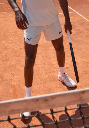 Tennis shorts in light gray with black logo, paired with white athletic shoes and socks. Player holds a tennis racket on clay court.