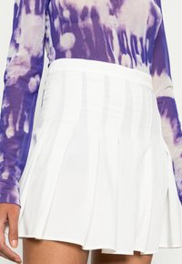 Saia mini plissada branca com uma textura suave e uma cinta ajustada, emparelhada com uma blusa de mangas longas em tie-dye roxa.