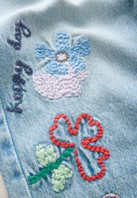 Jasnoniebieski denim z kolorowymi haftowanymi wzorami kwiatowymi: niebieski kwiat, różowe akcenty oraz czerwonokształtny serce z zielonymi liśćmi.