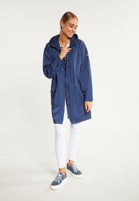 Jachetă impermeabilă de marină cu glugă, fermoar frontal și două buzunare frontale, purtată peste colanți albi și pantofi albaștri. Are un design casual.
