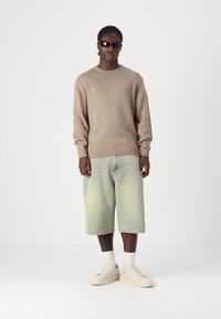 Beige Strickpullover, lockere hellblaue Denim-Shorts, weiße Sneakers mit strukturierter Sohle und dunkle Sonnenbrille. Schlichter Hintergrund.