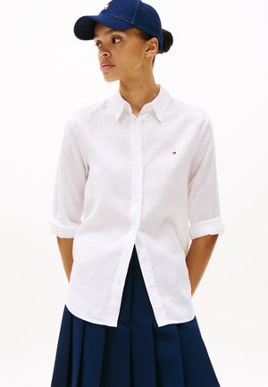 Femeie purtând o cămașă albă cu nasturi și mâneci suflecate, o fustă plisată bleumarin și o șapcă bleumarin, uitându-se în lateral.