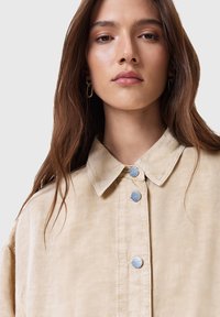 Camisa beige de botones con cuello, que cuenta con botones de metal azul y una tela texturada. Caída suave y corte suelto.