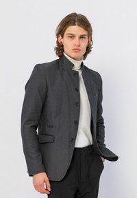 Donkergrijze op maat gemaakte blazer met een mandarin kraag, een sluiting met knopen aan de voorkant en zijzakken; gecombineerd met een lichte coltrui en zwarte broek.