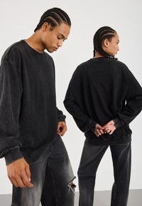 Sorte oversized hettegensere med slitt tekstur og matchende jeans, med ribbestrikkede mansjetter og en avslappet passform. Bakgrunnen er ensfarget hvit.