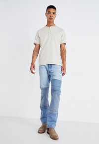 Beige pruhovaný tričko s krátkym rukávom henley s tromi gombíkmi, spárované so svetlomodrými patchwork džínsy a béžovými členkovými botami.