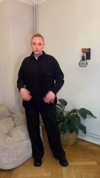 Sort skjorte med knapper og afslappet pasform, kombineret med løstsiddende sorte bukser og sorte sko, mod en minimalistisk indretning med planter.