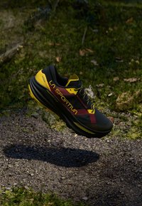 Melnas un dzeltenas La Sportiva taku skriešanas kurpes, kas plūst virs akmeņainas un netīras meža takas ar zaļu lapu fonu.