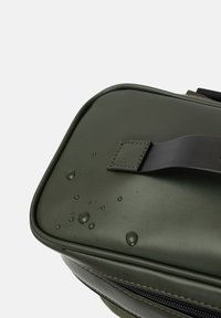 Sac étanche vert, forme rectangulaire, texture lisse, avec une sangle noire et des gouttes d'eau éparpillées sur la surface.