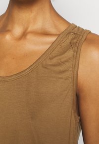 Detailní záběr horní části hrudníku a ramene v hnědém topu bez rukávů s viditelným klíční kostí a hladkou kůží.