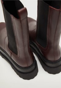 Bottines en cuir marron avec des côtés élastiques noirs, semelles épaisses noires et coutures détaillées le long des bords. Design à bout arrondi.