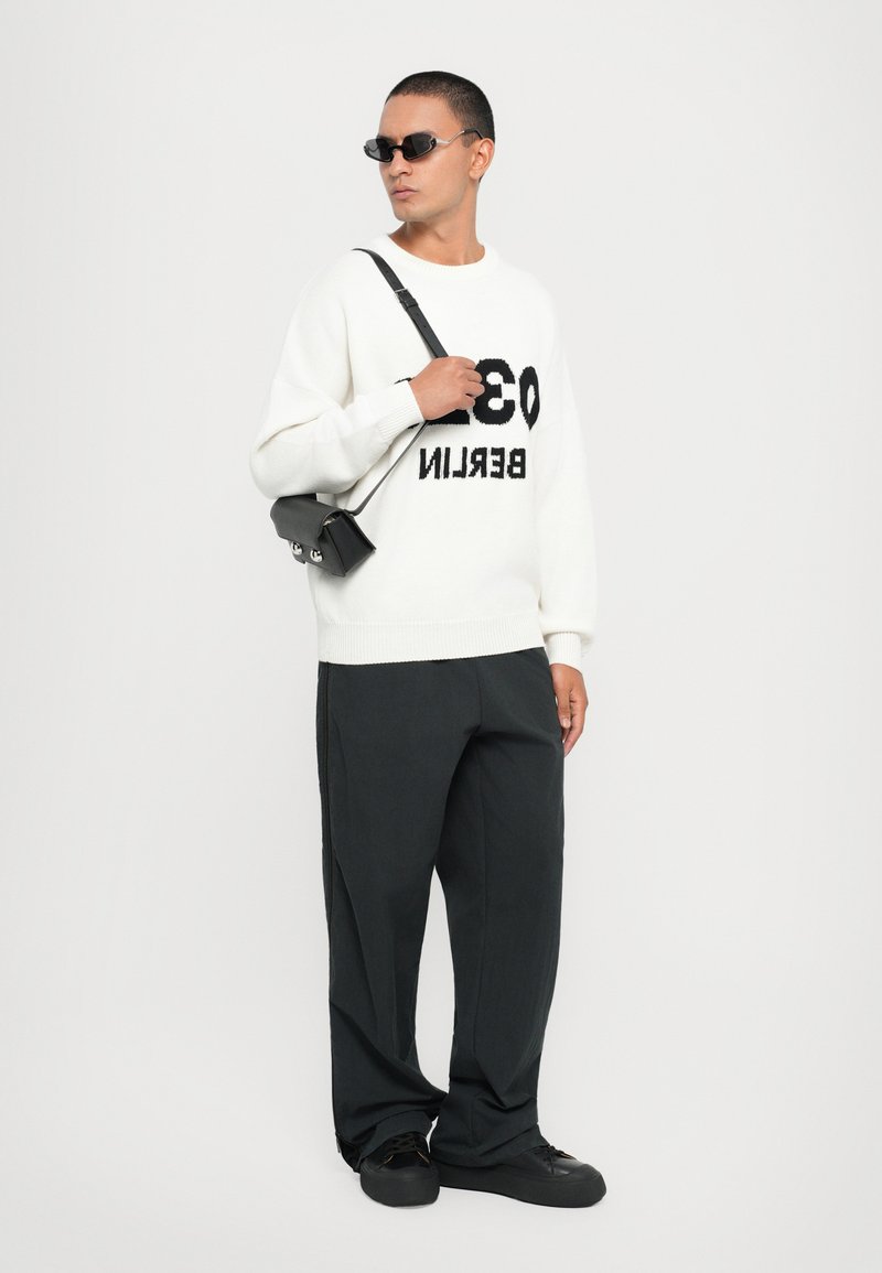 Weißer Strickpullover mit schwarzem "BERLIN 03"-Text, kombiniert mit schwarzen weit geschnittenen Hosen und schwarzen Sneakers. Modell trägt getönte Sonnenbrille und Umhängetasche.