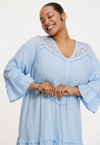 Hellblaues, leinenartiges Kleid mit V-Ausschnitt, Rüschensäumen, Knopfleiste und durchscheinendem, gemustertem Oberteil. Lässiger, fließender Stil.