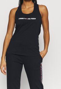 Czarny top na ramiączkach z okrągłym dekoltem, wyposażony w biało-kolorowe logo "TOMMY HILFIGER". Gładka tkanina i dopasowany fason, zestawione z czarnymi spodniami.