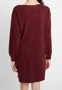 Abito lungo in maglia bordeaux con vestibilità rilassata, maniche oversize, dettagli a costine sul collo e spacchi laterali per facilitare i movimenti.