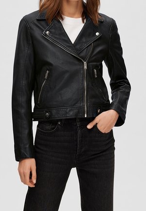 Femme portant une veste de motard en cuir noir avec des fermetures éclair et des boutons argentés, associée à un jean noir et une chemise blanche.