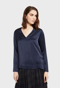 Saka plava satenska bluzica s V-izrezom i dugim rukavima, koja se ističe suptilnim sjajem, ravnim krojem i bočnim vezicama za prilagodivu veličinu.