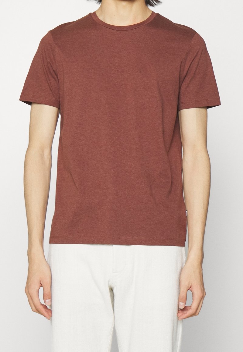 Personne portant un t-shirt marron uni à manches courtes et un pantalon blanc, debout devant un fond clair uni.