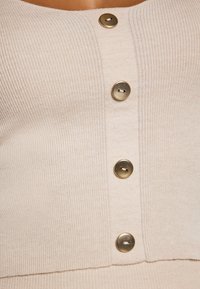 Beige ribbattu lõngast ülikond, millel on sügava kaelusega kujundus ja viis kuldset nööpi ees. Sile tekstuur ja keha järgi tihendatud disain.