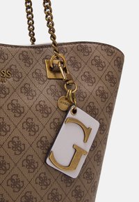 Brun tygkasse med guldkedja som axelrem, präglat logomönster och en vit etikett med en gulddetalj i form av ett "G".