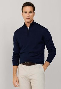 Muž v tmavomodrom svetri so štvrťzipsom, béžových nohaviciach a hnedom tkaničkovom opasku, stojaci s jednou rukou v vrecku na jednoliatom pozadí.