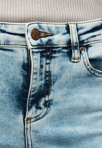 Φαρδιά τζιν από acid-washed τζην σε ανοιχτό μπλε χρώμα με μεταλλικό κουμπί και θηλιά ζώνης στη μέση, συνδυασμένα με μπεζ ριμπ τοπ που είναι μπουσουλιασμένο μέσα.