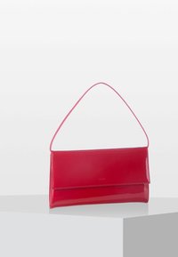 Bolso rectangular rojo brillante con una sola correa delgada exhibido en un pedestal blanco sobre un fondo gris claro liso.