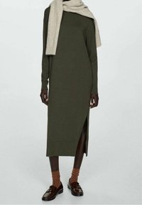 Robe en maille vert olive à longueur genou avec manches longues et fente latérale. Portée avec une écharpe beige, des chaussettes côtelées marron et des mocassins foncés.