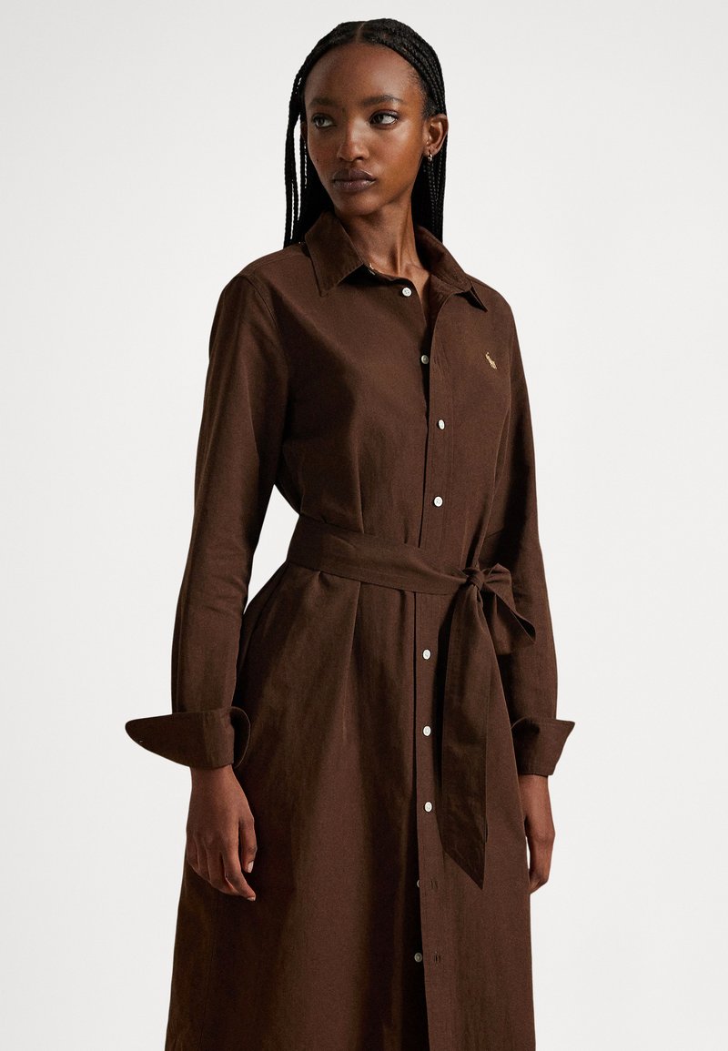 Woman with braided hair wearing a long-sleeve, buttoned brown dress with a tied waist belt, looking slightly to her left against a plain background.