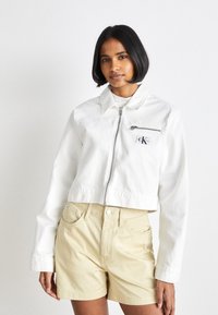 Giacca bianca corta con zip frontale, colletto e tasca sul petto; indossata con pantaloni corti beige chiaro. Tessuto in cotone liscio.