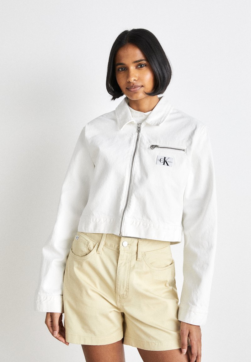 Giacca bianca corta con zip frontale, colletto e tasca sul petto; indossata con pantaloni corti beige chiaro. Tessuto in cotone liscio.