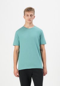 Tricou din bumbac de culoare teal deschis, cu guler rotund și mâneci scurte, având o croială lejeră și o textură fină. Purtați cu pantaloni negri.