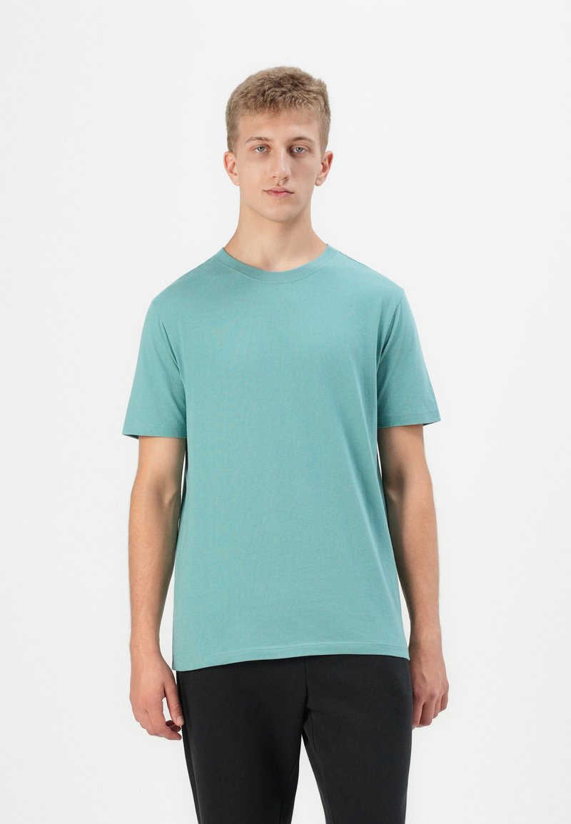 Tricou din bumbac de culoare teal deschis, cu guler rotund și mâneci scurte, având o croială lejeră și o textură fină. Purtați cu pantaloni negri.