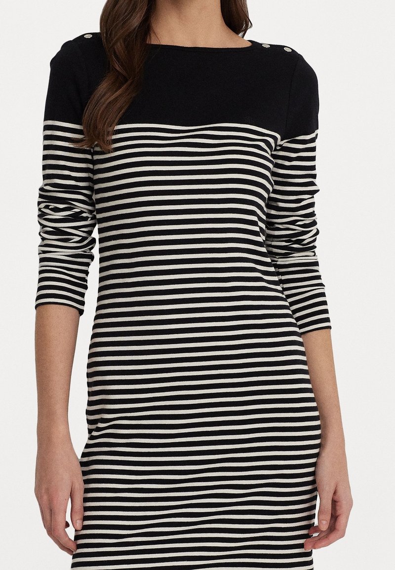 Femme portant une robe à manches longues rayée horizontalement en noir et blanc, avec des épaules unies noires et des détails de boutons.